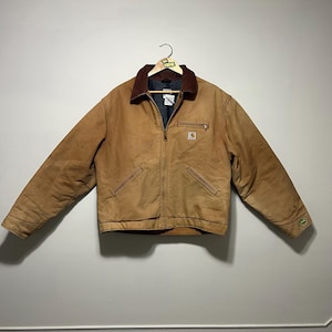 Puede incluir: Chaqueta de trabajo Carhartt marr&oacute;n con cuello de pana, cremallera y m&uacute;ltiples bolsillos. La chaqueta tiene un peque&ntilde;o logotipo en el pecho y un logotipo verde y blanco en la manga. La chaqueta est&aacute; colgada en una percha.