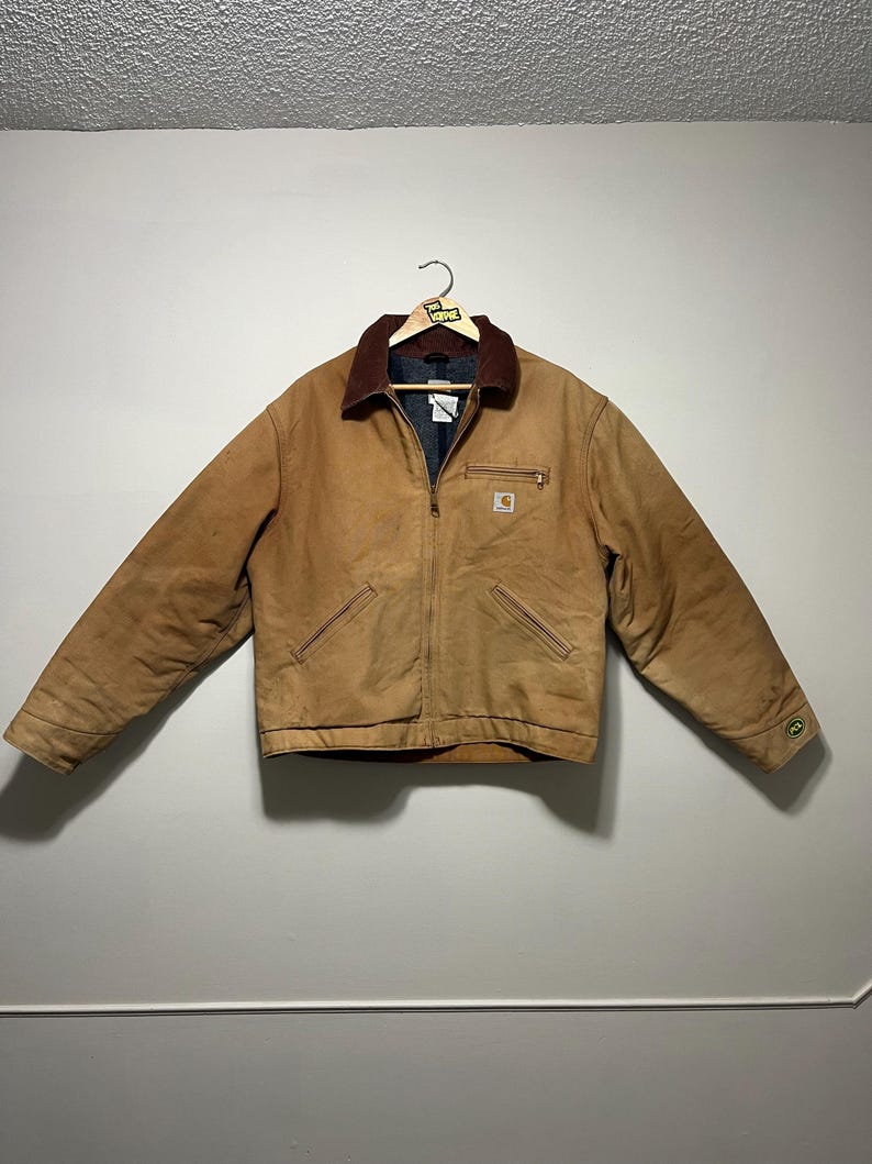 Puede incluir: Chaqueta de trabajo Carhartt marr&oacute;n con cuello de pana, cremallera y m&uacute;ltiples bolsillos. La chaqueta tiene un peque&ntilde;o logotipo en el pecho y un logotipo verde y blanco en la manga. La chaqueta est&aacute; colgada en una percha.