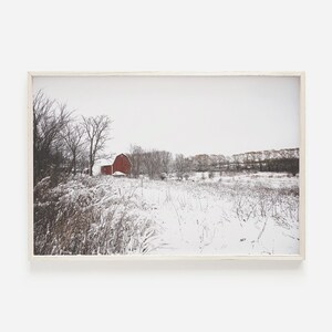 May include: A red barn stands in a snowy field with trees in the background. The snow covers the ground and the trees. The sky is a light gray.