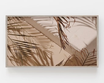 Puede incluir: Una fotografía enmarcada de una pared blanca con sombras proyectadas por hojas de palmera. Las hojas son marrones y secas, y las sombras son largas y delgadas. La fotografía está en un marco de madera claro.