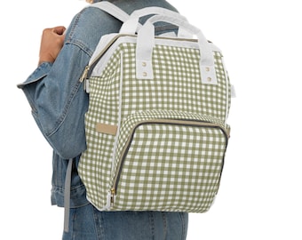 Puede incluir: Una mochila de pañales de estilo mochila con estampado de cuadros verdes y blancos con detalles dorados. La mochila tiene un compartimento principal grande, un bolsillo frontal más pequeño y dos bolsillos laterales.