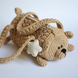 May include: A tan crocheted teddy bear-shaped bag with a matching strap and handles. The bag features a small, off-white star-shaped charm attached with a silver clasp. The bear has black eyes and a black nose.