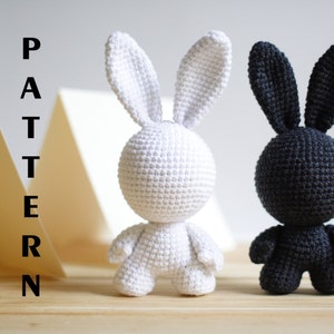 May include: Two crochet bunny figures, one white and one black, stand side-by-side. The bunnies have large ears and small arms and legs. The word "PATTERN" is written vertically in black letters on the left side of the image.
