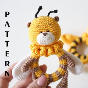 May include: A crocheted bee rattle toy with a yellow and brown striped body, white wings, and a yellow and brown striped ring. The bee has a white face with black eyes and a brown nose.