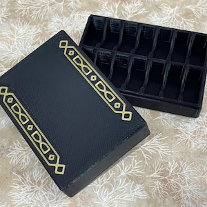 May include: A black rectangular box with a hinged lid, open to reveal a compartmented interior. The lid is decorated with gold geometric designs. The box is likely for storing small items, such as jewelry or game pieces.