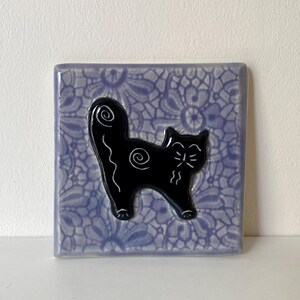 May include: A square ceramic tile featuring a light purple background with a dark purple lace-like floral pattern. Centered on the tile is a glossy black cat silhouette with white line details, including a spiral on its body and tail.
