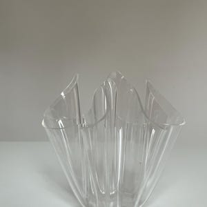 May include: A clear, decorative vase with a unique, ruffled design. The vase is made of transparent material, allowing light to pass through. The shape is reminiscent of a flower or a folded fabric, adding a touch of elegance to any space.
