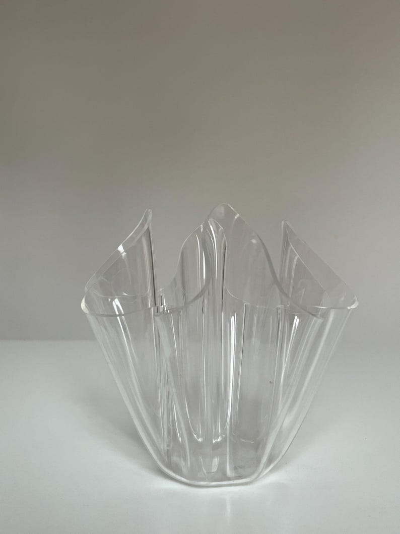 May include: A clear, decorative vase with a unique, ruffled design. The vase is made of transparent material, allowing light to pass through. The shape is reminiscent of a flower or a folded fabric, adding a touch of elegance to any space.