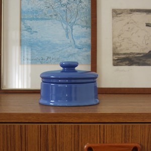 Pode incluir: Um pequeno pote de cerâmica azul brilhante com tampa e um puxador simples em forma de botão. O recipiente possui um corpo cilíndrico com uma base ligeiramente alargada, apresentando uma cor azul sólida e uniforme em toda a peça.