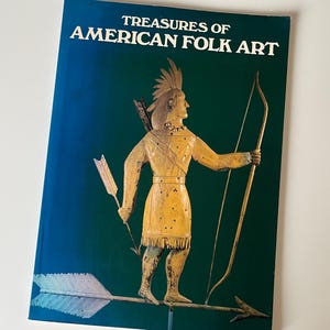 Może przedstawiać: Książka zatytułowana "Treasures of American Folk Art" z turkusową okładką. Na okładce znajduje się żółta metalowa figura Indianina z łukiem i strzałą. Tło jest w kolorze ciemnozielonym.