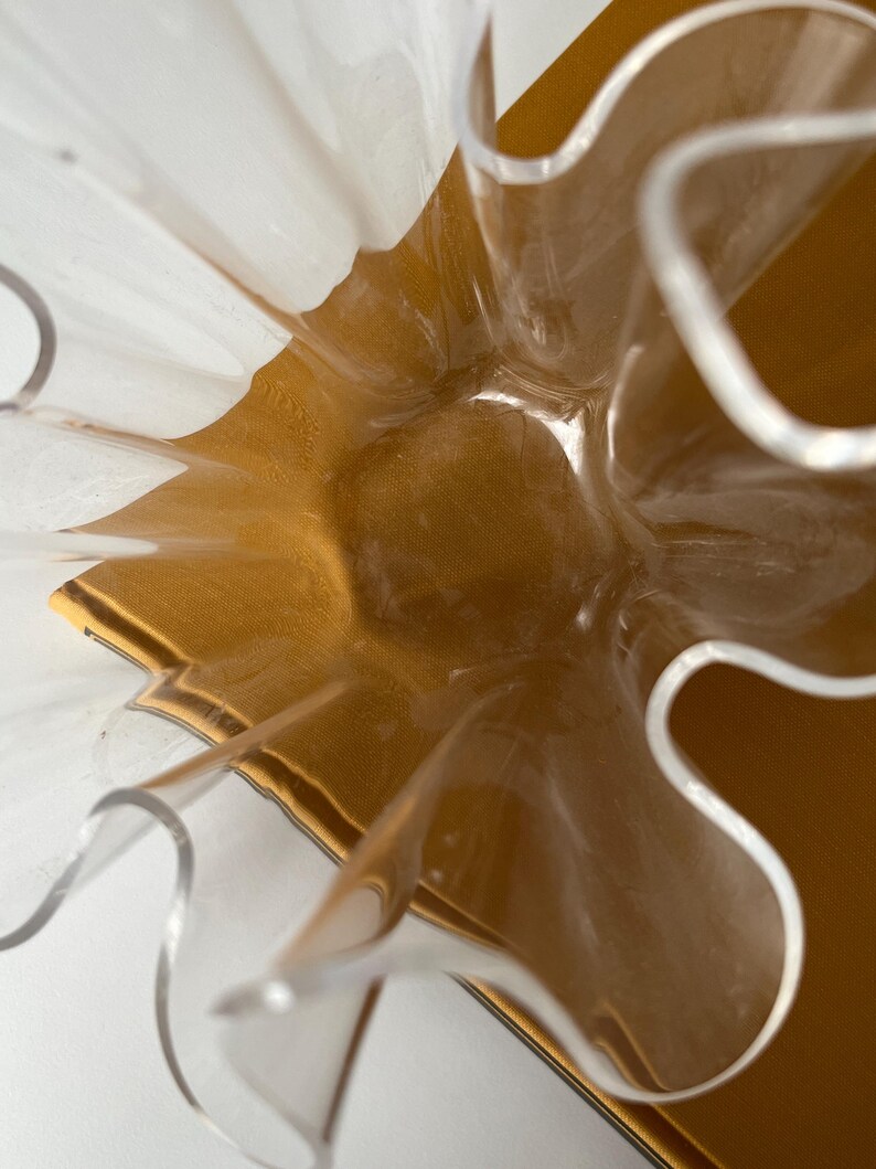 May include: A clear, wavy-edged decorative bowl with a gold-colored interior. The bowl's design features a series of curved, petal-like shapes, creating a unique and artistic appearance. The bowl is set against a white background.