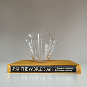 May include: A clear, fluted glass bowl sits atop a gold-colored book titled "THE WORLD'S ART 2". The book's spine displays the text "DENIS & DE VRIES" and "COLLINS". The bowl has a unique, wave-like design.