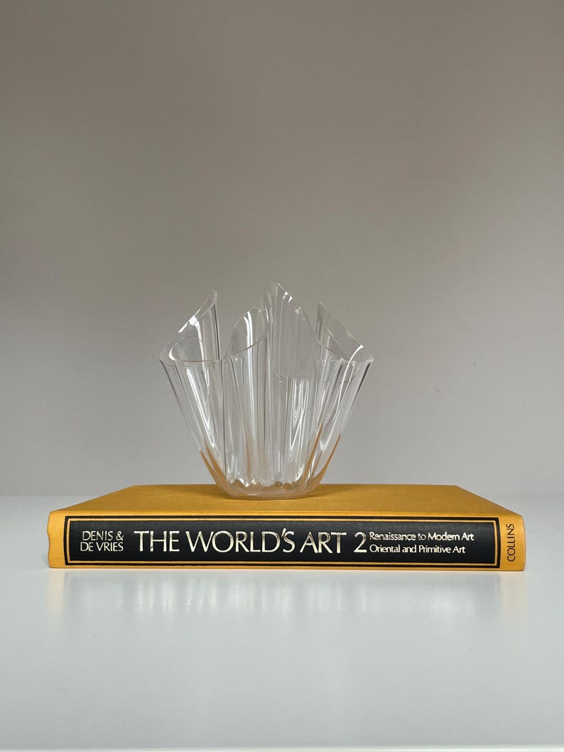 May include: A clear, fluted glass bowl sits atop a gold-colored book titled "THE WORLD'S ART 2". The book's spine displays the text "DENIS & DE VRIES" and "COLLINS". The bowl has a unique, wave-like design.