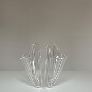 May include: A clear, decorative glass vase with a unique, folded design. The vase has a square base and a ruffled, open top. The transparent material allows light to pass through, highlighting the intricate folds and curves of the glass.