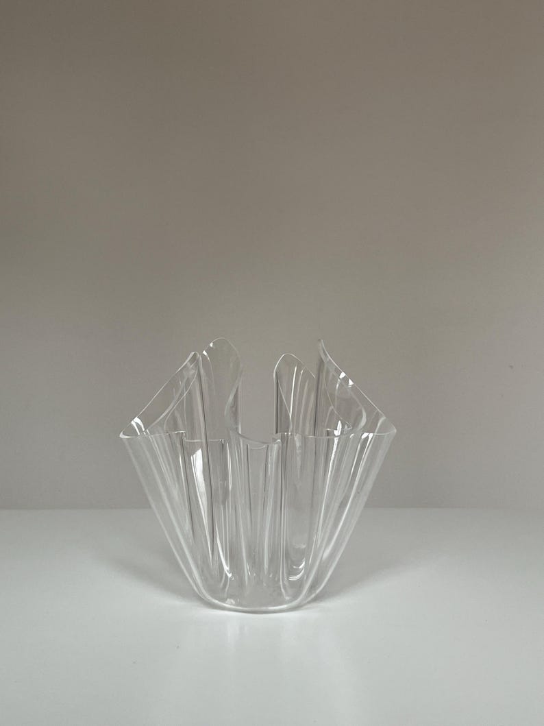 May include: A clear, decorative glass vase with a unique, folded design. The vase has a square base and a ruffled, open top. The transparent material allows light to pass through, highlighting the intricate folds and curves of the glass.