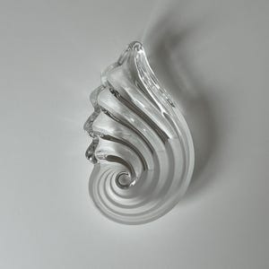 Könnte beinhalten: Eine Skulptur aus klarem Glas in Form eines Nautilus-Gehäuses mit einem spiralförmigen Design aus abwechselnd transparenten und mattierten Glasrippen, die sich zu einer Spitze hin wölben und eine texturierte, reflektierende Oberfläche bilden.