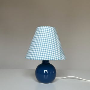 May include: A navy blue ceramic table lamp with a round base and a light blue and white gingham lampshade. The lamp has a white power cord. The lamp is on a white surface.