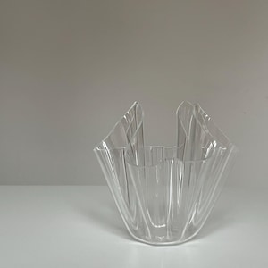 May include: A clear, decorative glass bowl with a unique, folded design. The bowl has a flared, ruffled top and a wide, open interior. The transparent material allows light to pass through, creating interesting shadows.