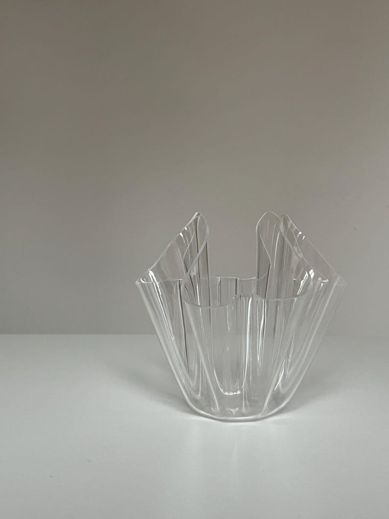 May include: A clear, decorative glass bowl with a unique, folded design. The bowl has a flared, ruffled top and a wide, open interior. The transparent material allows light to pass through, creating interesting shadows.