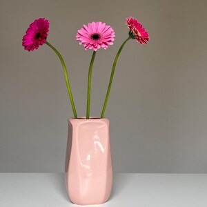 Könnte beinhalten: Eine rosa Keramikvase mit drei Gerbera-Blüten. Die Blüten haben rosa Blütenblätter und dunkle Zentren. Die Vase ist hellrosa und hat eine geometrische Form. Die Stiele der Blumen sind grün.