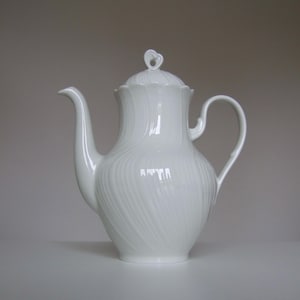 Op de afbeelding: Een witte porseleinen theepot met een glanzende afwerking en een subtiel, wervelend reliëfpatroon over het lichaam. De theepot heeft een gebogen tuit, een C-vormig handvat en een bijpassend deksel met een kleine, hartvormige knop.