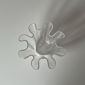May include: A clear, decorative glass bowl with a unique, wavy, petal-like rim. The bowl's design features a series of curved edges, creating a flower-like shape. The transparent glass allows light to pass through, highlighting its form.