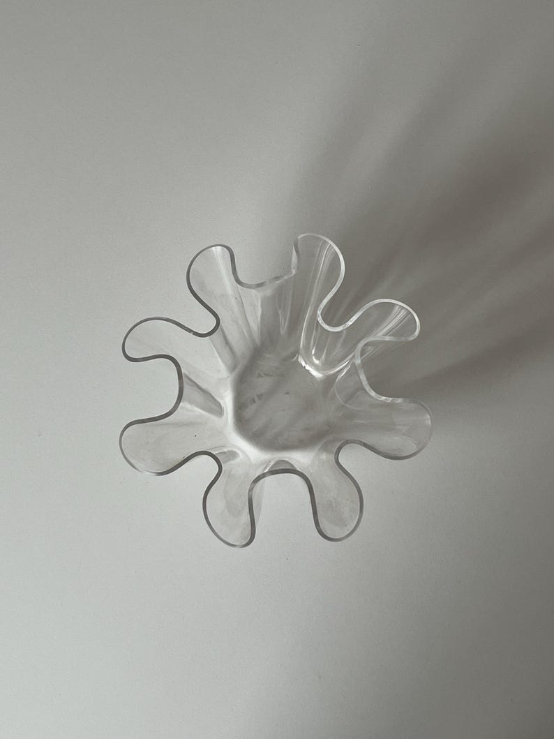 May include: A clear, decorative glass bowl with a unique, wavy, petal-like rim. The bowl's design features a series of curved edges, creating a flower-like shape. The transparent glass allows light to pass through, highlighting its form.