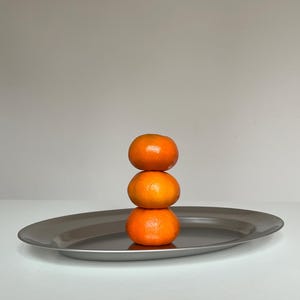 Op de afbeelding: Drie feloranje mandarijnen gestapeld op een zilveren ovaal serveerblad. De lade heeft een glad, reflecterend oppervlak en een licht verhoogde rand. De achtergrond is effen lichtgrijs.