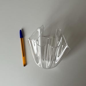 May include: A clear, wavy-edged decorative bowl. The bowl is made of transparent plastic and has a unique, folded design. A yellow and blue pen is next to the bowl for scale.