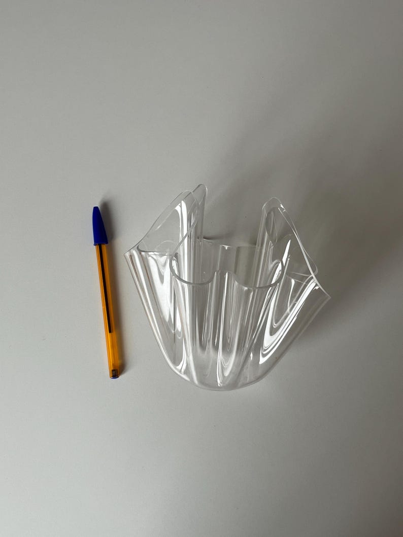 May include: A clear, wavy-edged decorative bowl. The bowl is made of transparent plastic and has a unique, folded design. A yellow and blue pen is next to the bowl for scale.