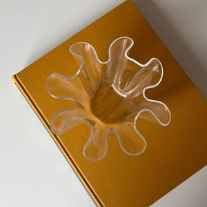 May include: A clear, petal-shaped glass bowl sits atop a gold-colored book. The bowl has a unique, wavy design, with multiple curved edges. The book's cover has a smooth, reflective surface.