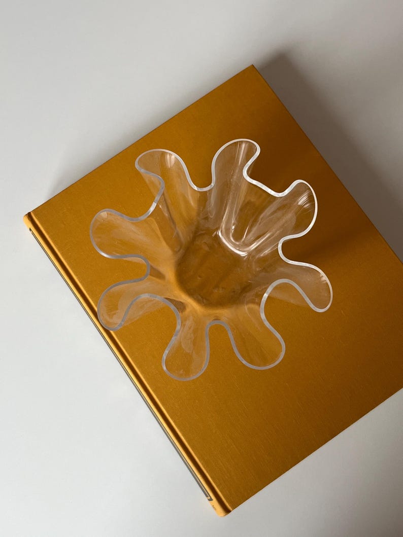 May include: A clear, petal-shaped glass bowl sits atop a gold-colored book. The bowl has a unique, wavy design, with multiple curved edges. The book's cover has a smooth, reflective surface.