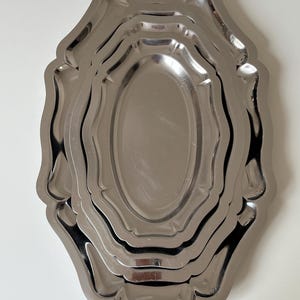 Peut inclure: Un ensemble de trois plateaux en métal argenté imbriqués avec une finition polie et réfléchissante. Chaque plateau présente une forme ovale allongée avec un bord festonné décoratif. Les plateaux sont empilés les uns dans les autres.