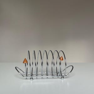 May include: A chrome-plated metal toast rack featuring a series of six vertical wire loops for holding slices of bread. The rack includes two small, pointed wooden finials on the outer loops and rests on four small, spherical metal feet.