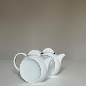 Può includere: Due teiere in ceramica bianca. Una teiera ha un grande design circolare sul lato, un manico curvo e un coperchio. L'altra teiera ha un bordo blu attorno al coperchio. Entrambe hanno un beccuccio.