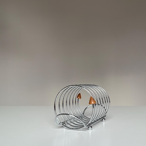 May include: A chrome-finished metal wire rack featuring a series of concentric circular loops. The structure is supported by small spherical feet and includes two small, pointed, light brown wooden accents attached to the interior wire frame.