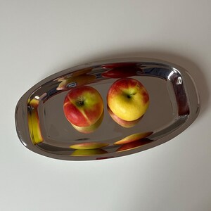 Op de afbeelding: Een ovale serveerschaal van gepolijst roestvrij staal met twee appels. De appels zijn rood en geel, met een glanzend oppervlak. De schaal weerspiegelt de appels en de omgeving.
