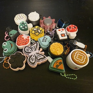 May include: A collection of colorful, decorative keychains in various shapes, including a strawberry, a frog, a butterfly, and a coffee cup. The keychains are made of plastic and have metal chain attachments.