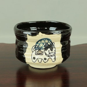 May include: A black and white ceramic teacup featuring a hand-painted design of a cat wearing a blue and white patterned hat. The cup has a black rim and a white interior.