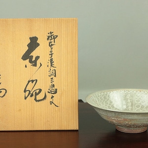 May include: A wooden box with Japanese calligraphy on the front, next to a white ceramic bowl with a decorative pattern. The bowl has a light brown glaze and a slightly raised rim.