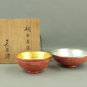 Op de afbeelding: Twee rode keramische kommen met gouden en zilveren accenten. De kommen bevinden zich in een houten doos met Japanse karakters op de deksel.