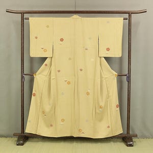 以下が含まれることがあります： 薄い黄色の着物、花柄の刺繍入り。濃い茶色の木製ラックに展示されています。着物には、赤、オレンジ、銀色の花模様が散りばめられています。袖が広げられ、衣服のデザインと質感が示されています。
