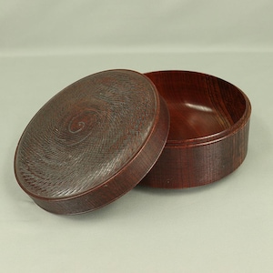 May include: A round wooden box with a lid. The lid has a spiral design carved into it. The box is made of dark wood and has a reddish-brown hue.