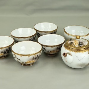 May include: A white porcelain tea set featuring a teapot with a side handle and five matching tea bowls. Each piece is decorated with a landscape scene in muted tones and finished with a decorative gold rim along the top edges of the vessels.