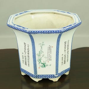 May include: An octagonal white ceramic planter with blue and white geometric patterns around the rim. The planter has a green and black floral design with Chinese calligraphy on the sides. The calligraphy reads: "三月王所寒春兰西丝丝不有千數得卖花声到了一种清奇胜"