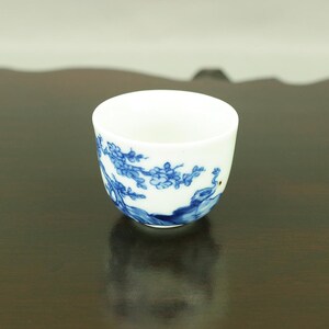 Peut inclure: Une petite tasse à thé en céramique blanche avec un motif floral bleu et blanc. La tasse est posée sur une surface en bois brun foncé.
