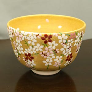 May include: A golden-yellow ceramic bowl featuring a detailed pattern of white, light pink, and dark red cherry blossom flowers across the exterior and interior rim. The bowl has a smooth, rounded shape and a small, circular white base at the bottom.