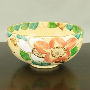 May include: A cream-colored ceramic bowl with a floral design. The bowl has a scalloped rim and is decorated with pink and white flowers, green leaves, and gold accents.
