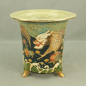 Könnte beinhalten: Eine dekorative Keramikvase mit cremefarbenem Rand und goldenen Akzenten. Die Vase zeigt eine detaillierte Illustration eines goldenen Drachen vor einem türkisfarbenen Hintergrund mit Wolken und floralen Mustern. Die Vase wird von drei goldfarbenen Füßen getragen.