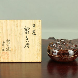 Puede incluir: Una pequeña caja redonda de cerámica marrón con un diseño en relieve de un dragón. La caja está en una caja de madera con escritura japonesa en la tapa.